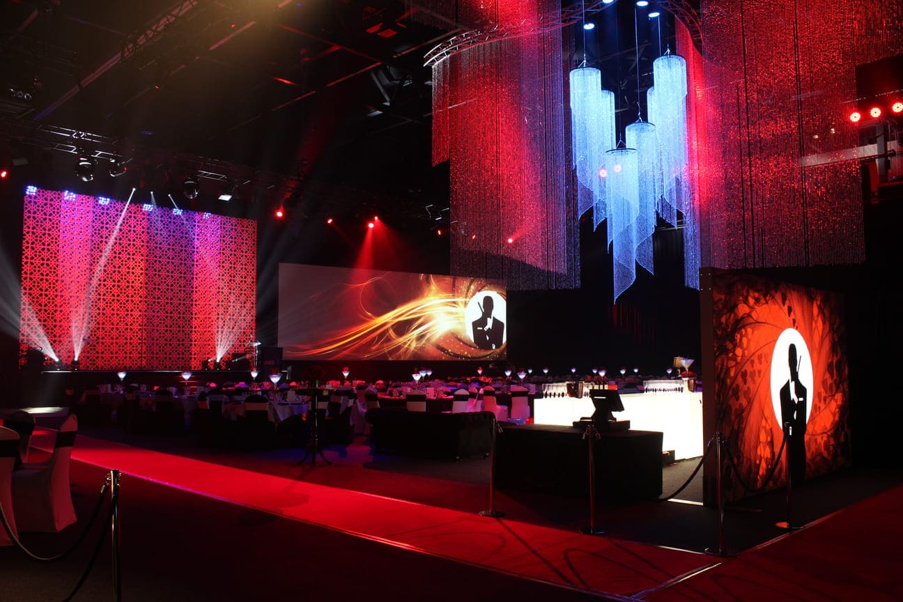 A glamorous event venue with dramatic lighting, large projection screens, round tables set for dining, and sparkling chandeliers. Red and purple hues create an elegant atmosphere, with a silhouette image on a lit panel.