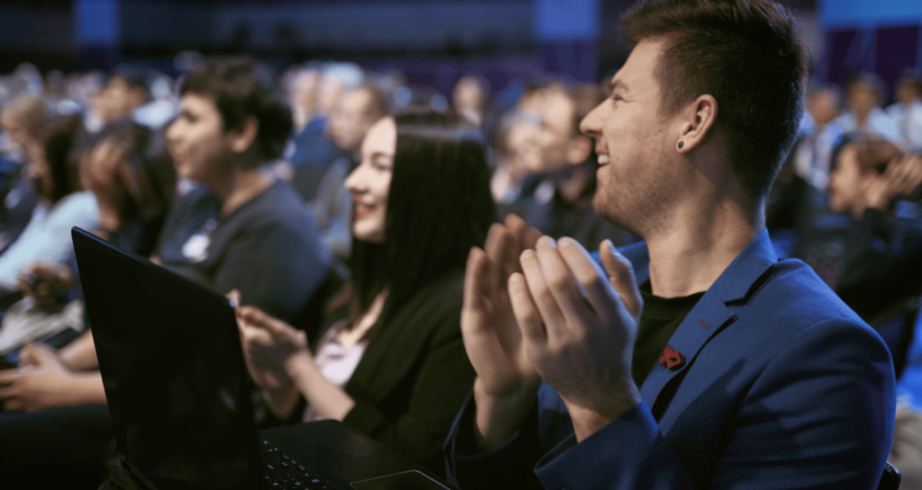 A group of people sitting in an audience, smiling and applauding. The focus is on a man in a blue blazer clapping, with a laptop on his lap. The background shows other attendees, blurred.