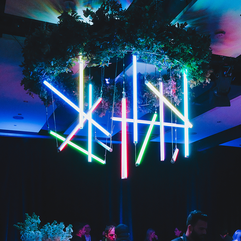 A modern chandelier with hanging neon lights in blue, green, and red, surrounded by leafy foliage, glows in a dark room at an event produced by a leading AV company Auckland, with people and plants visible in the background.