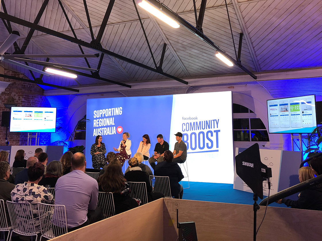 A panel of five people speaks onstage at a Facebook Community Boost roadshow event, with an audience seated in front. The stage displays “Supporting Regional Australia” as event screens show digital content. The venue has exposed beams and bright lighting.
