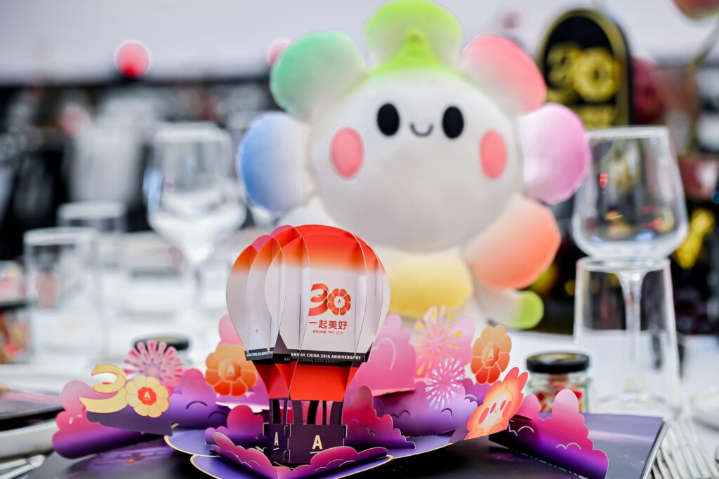 A colorful table display features a 3D pop-up of hot air balloons with 20 on them, surrounded by flowers. In the background, a plush toy with a rainbow-colored head and a smiling face is visible next to glassware.