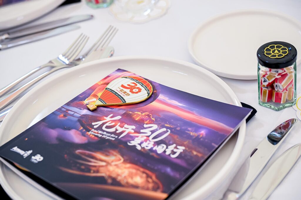 A place setting features a booklet with “30” and Chinese text on the cover, topped with a hot air balloon-shaped badge. A small jar of candy, plates, and cutlery are arranged neatly on a white tablecloth.