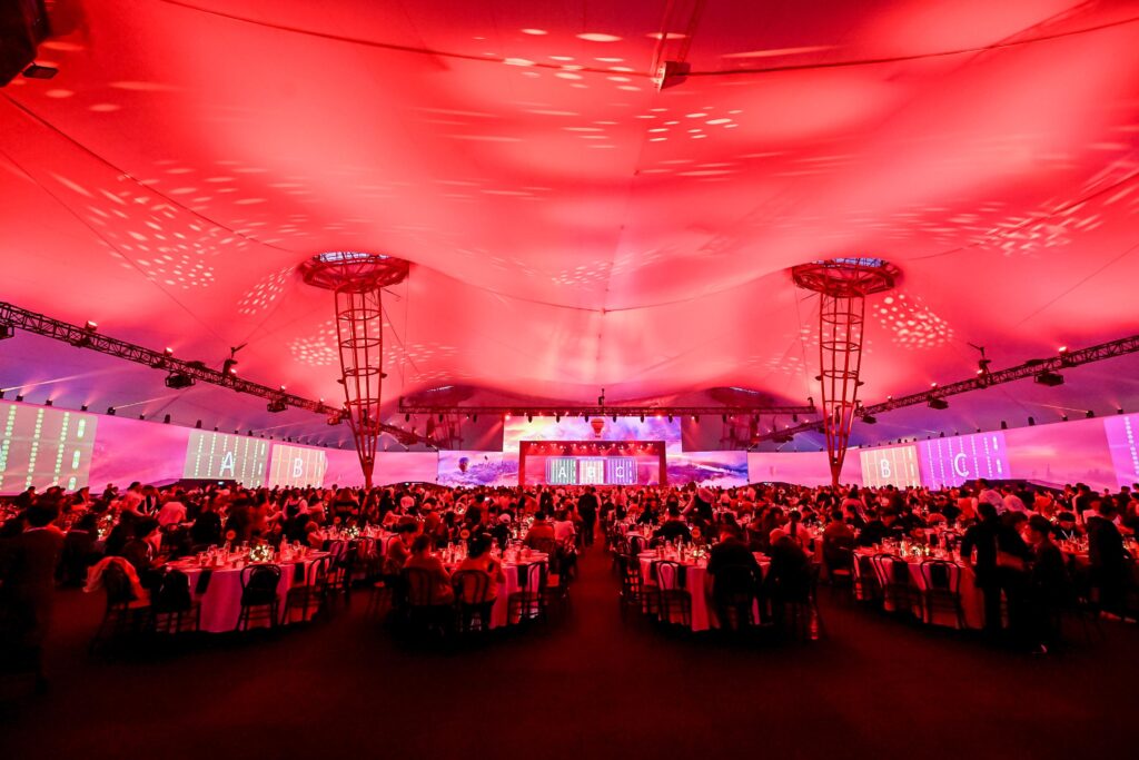 A large crowd sits at round banquet tables under a tent with dramatic red lighting, watching a stage at the front. The ceiling is illuminated with patterns, and lettered sections are visible on the tent walls.