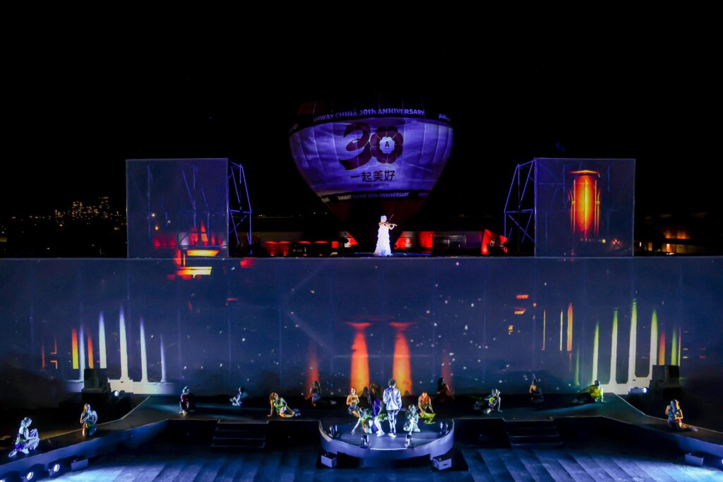 A stage performance at night with dancers in colorful costumes, a singer in white elevated above the stage, and a large hot air balloon in the background displaying 30th Anniversary and Chinese characters.