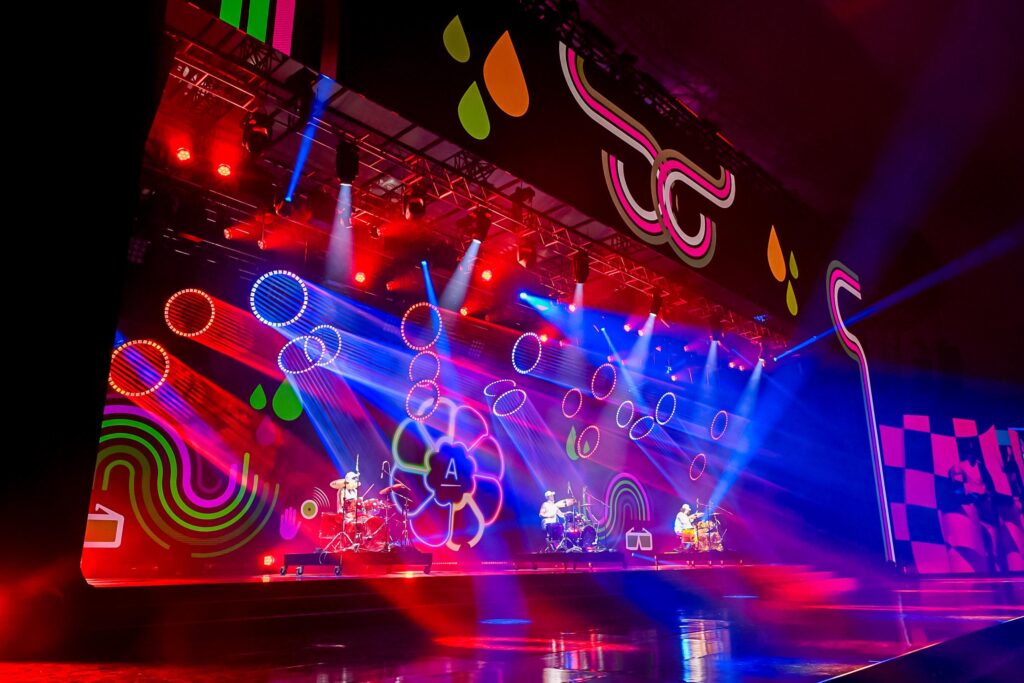 A vibrant concert stage with colorful lights, geometric patterns, and abstract shapes on the backdrop. Musicians perform under red, blue, and purple spotlights, creating a dynamic and energetic atmosphere.