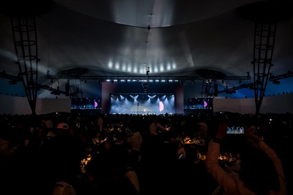 A large crowd in a dimly lit indoor venue faces a brightly lit stage with performers, under a high, curved ceiling. Some audience members are taking photos or videos.