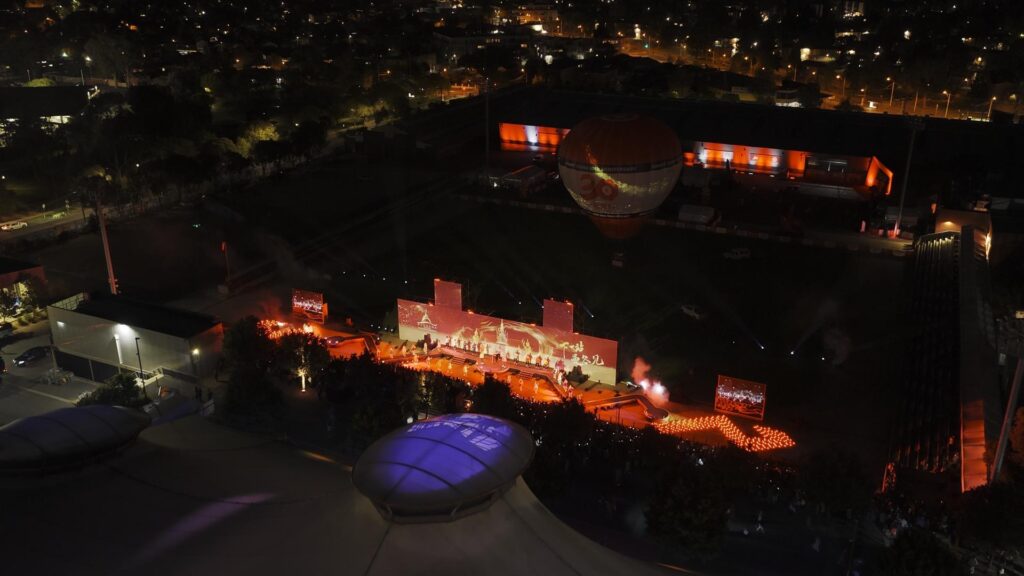An aerial night view of a stadium lit with red and orange lights, featuring a stage with performers, a crowd, and a large hot air balloon. City lights are visible in the background.