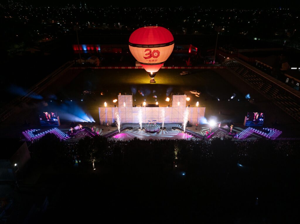 A brightly lit outdoor stage at night with colorful lights, an audience, and a large red and white hot air balloon displaying 30 floating above, viewed from above in a stadium setting.