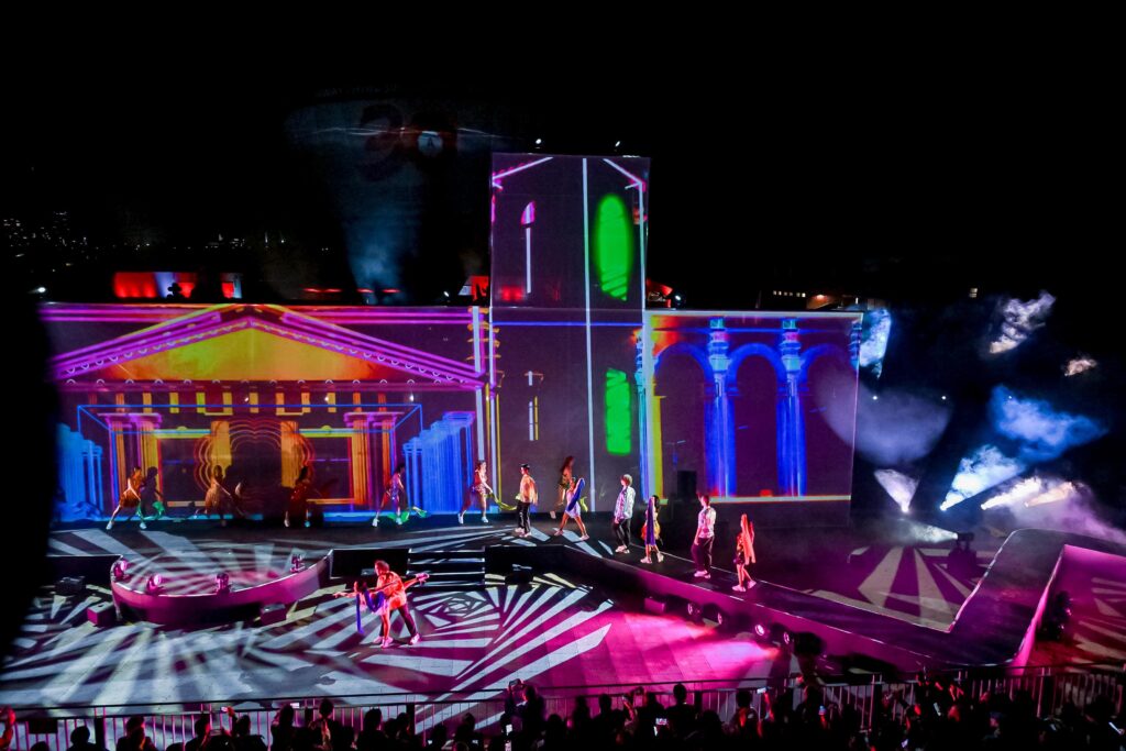 Colorful digital projections of classical architecture light up a stage at night, with performers walking on an elevated runway and smoke effects enhancing the dramatic scene.