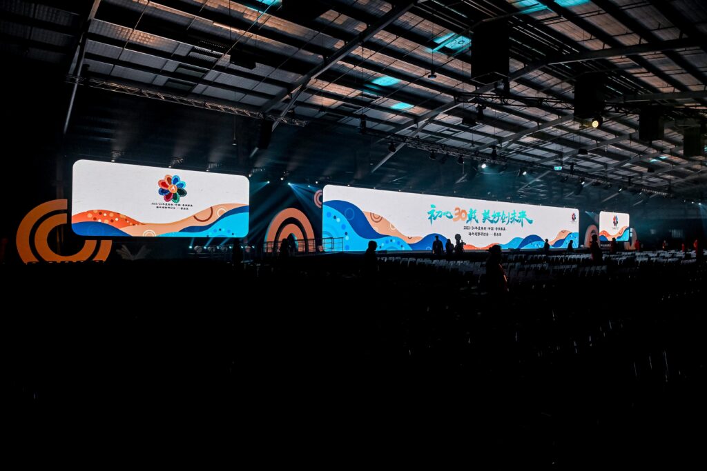 A large indoor event space with bright, colorful digital screens displaying Chinese text, graphics, and event logos. Rows of empty chairs fill the darkened foreground.