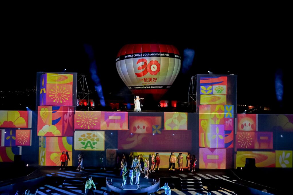 A colorful stage with vibrant, patterned projections features performers below and a person standing above. A large hot air balloon with 30th Anniversary is in the background, illuminated against the night sky.