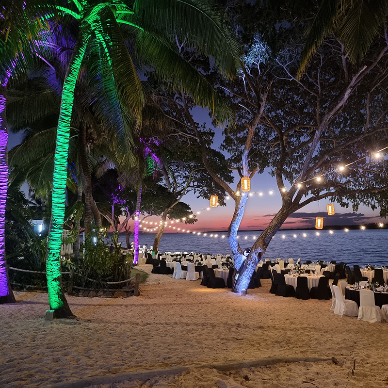 A beachside event setup at sunset by an event production company in Fiji, featuring dining tables with black and white cloths, string lights between palm trees, and trees illuminated by colorful purple and green lights.