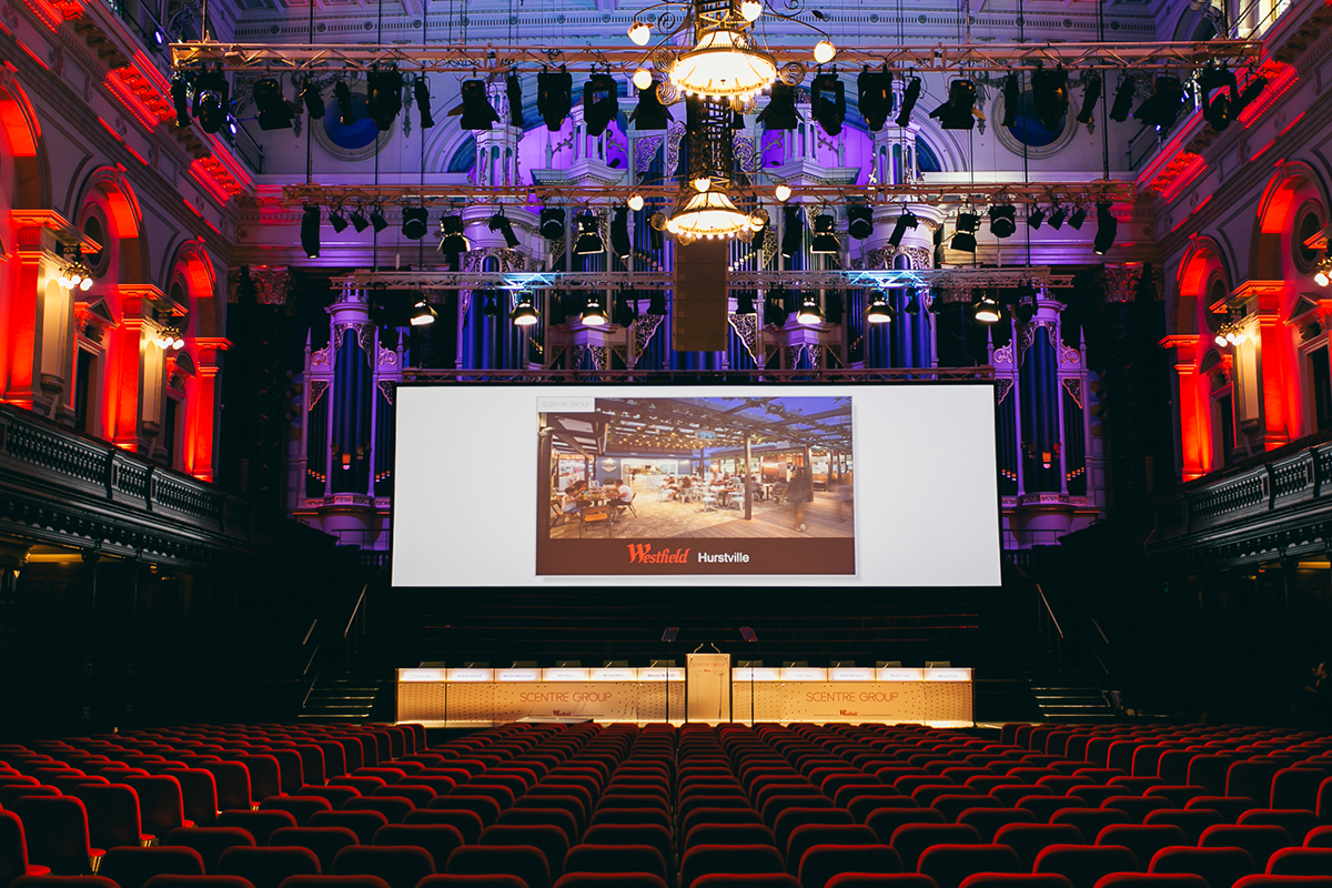 A grand, ornate auditorium with red seats faces a large screen displaying an image and the text Westfield Hurstville. Colorful lights illuminate the stage, high ceilings, and arched windows.