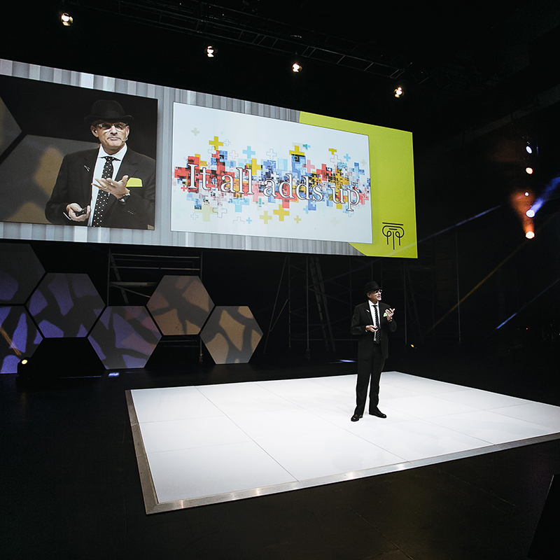 A man in a suit and hat speaks on a stage with geometric lighting. Behind him, a large screen displays his image and the word “Italiadadstop” over a colorful plus-sign pattern.
