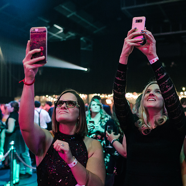 Two women at an indoor event hold up their phones, appearing to film or take photos. They are surrounded by a crowd under colorful lights. Both women wear black clothing and wristbands, and look focused on their screens.