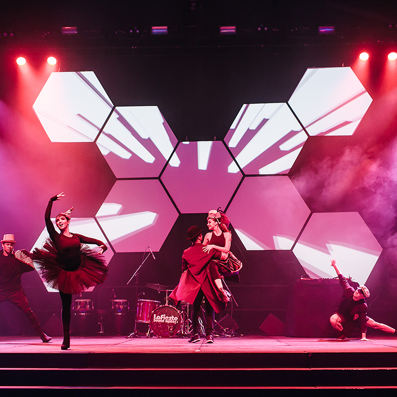 A group of dancers perform energetically on stage under dramatic red and purple lighting, with geometric hexagon screens displaying abstract visuals in the background.
