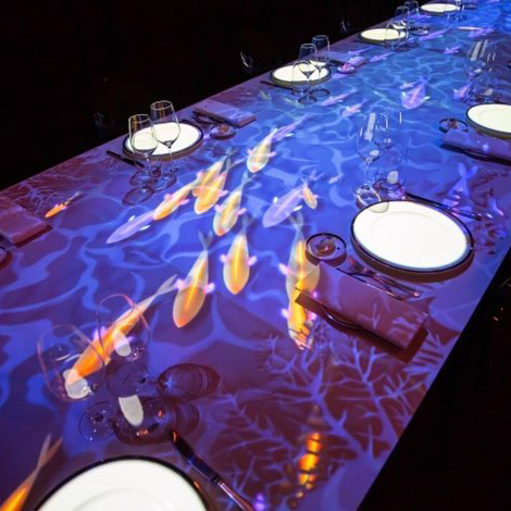 A long dining table is set with plates, glasses, and napkins, while a colorful light projection of swimming fish and water ripples decorates the tabletop, creating an underwater effect.