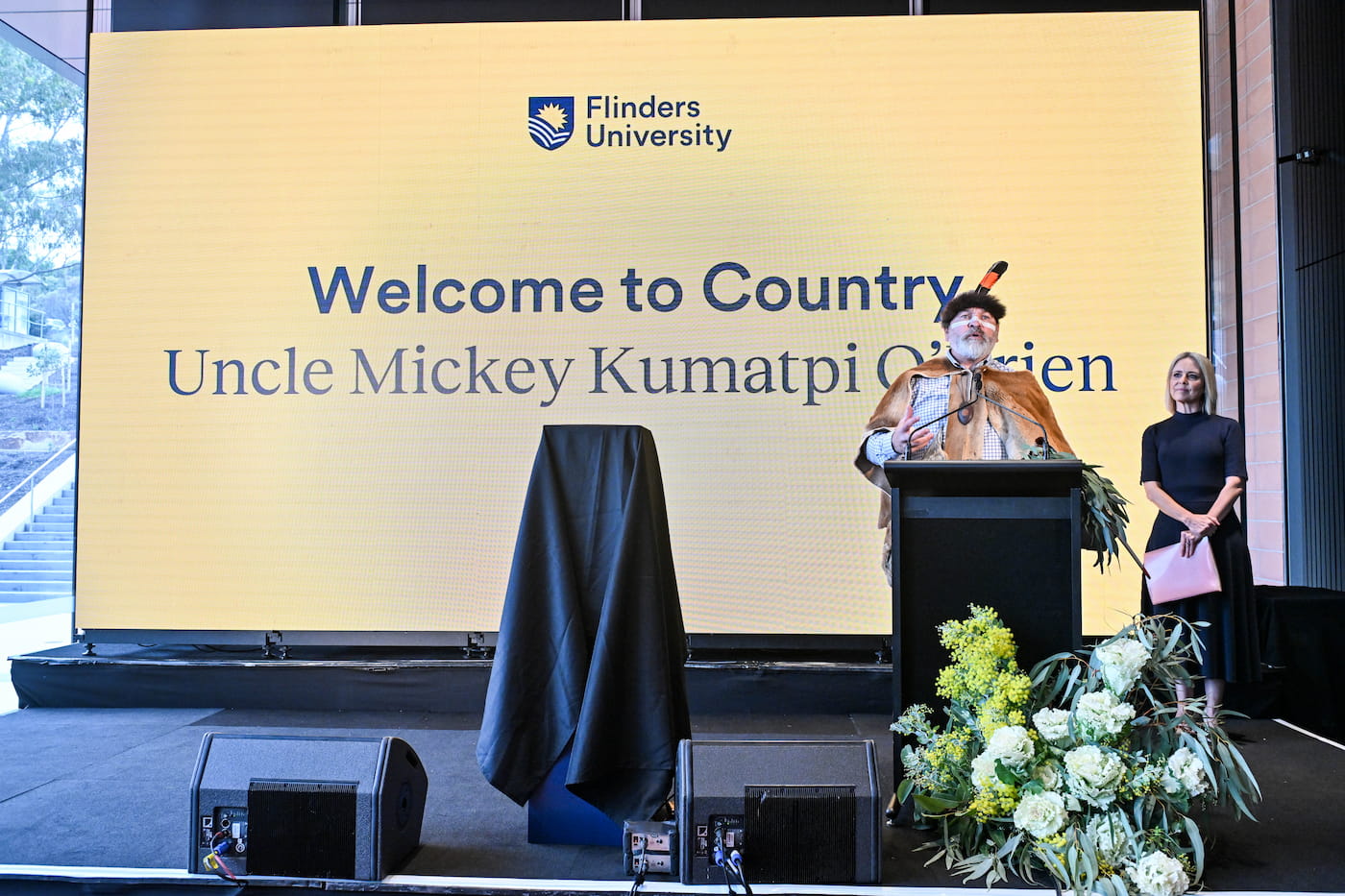 A man in traditional attire speaks at a podium on stage next to a woman, with a large screen behind them displaying Welcome to Country Uncle Mickey Kumatpi OBrien, Flinders University.