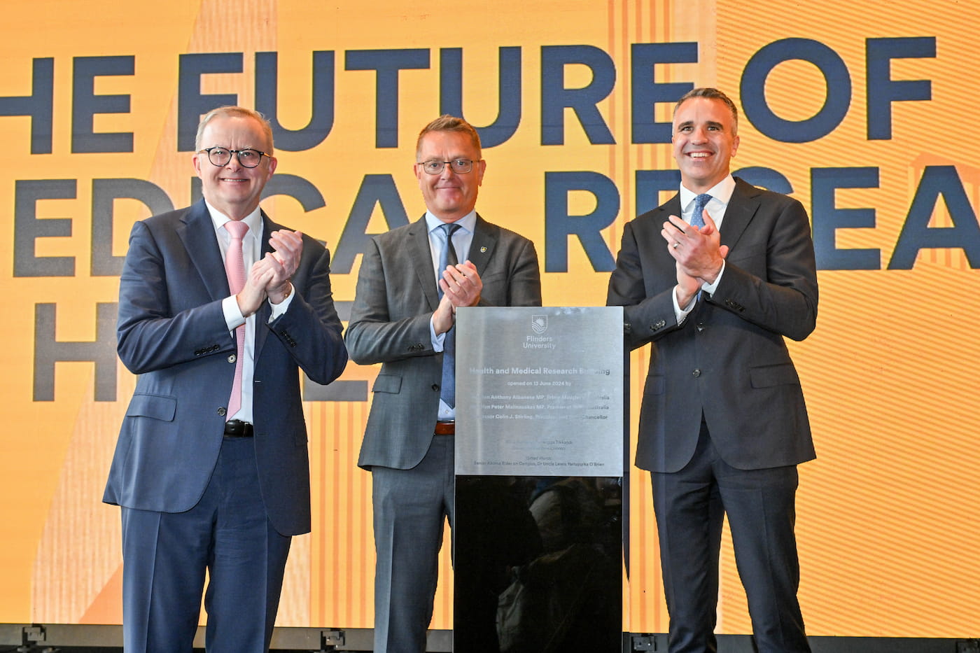Three men in suits stand smiling and applauding in front of a plaque, with a yellow background displaying the words “THE FUTURE OF MEDICAL RESEARCH.”.