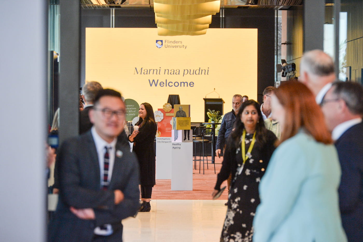 A group of people converse in a bright hallway; in the background, a large yellow sign with the Flinders University logo reads “Marni naa pudni Welcome.”.