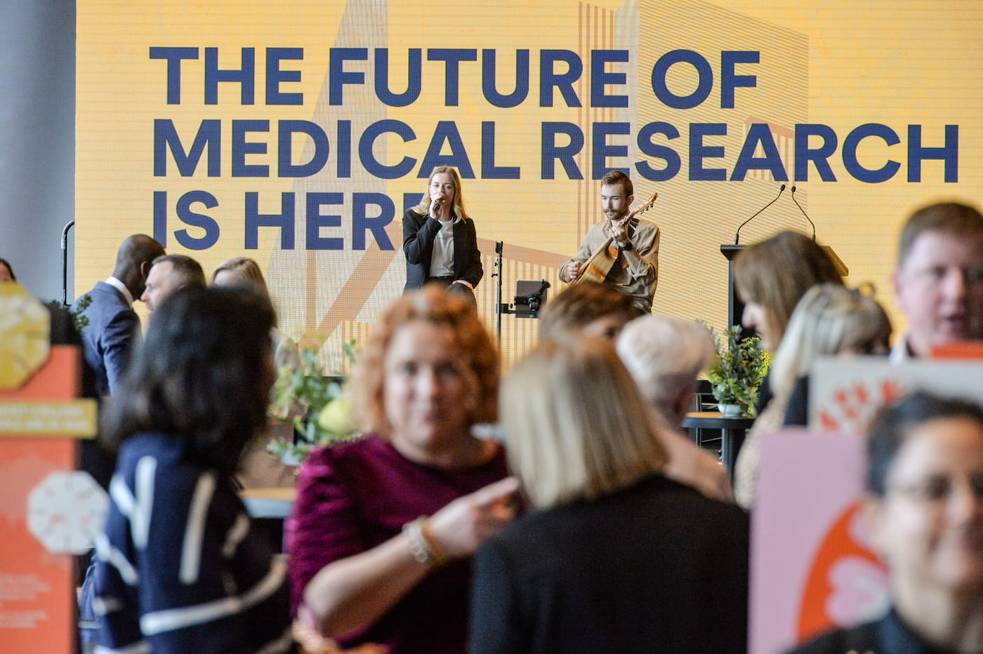 A crowd attends an event in front of a large yellow sign that reads, The future of medical research is here. Two musicians perform on stage in the background.