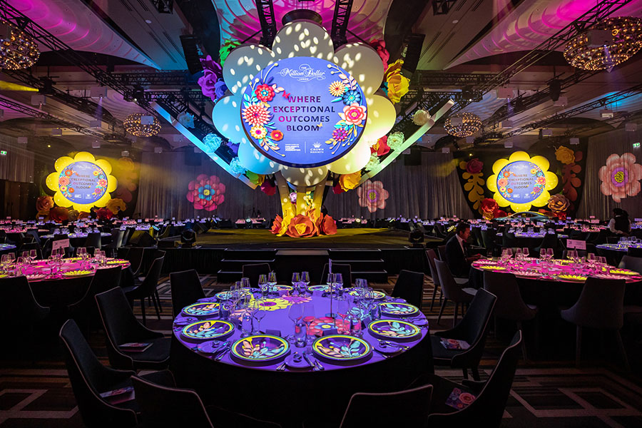A brightly lit event space decorated with colorful, oversized flowers and round tables set for dinner showcases different event types. A large sign on stage reads Where Exceptional Outcomes Bloom. The atmosphere is festive and vibrant.