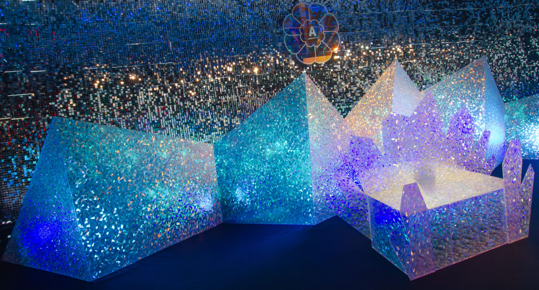 An art installation features large, iridescent geometric shapes resembling crystals or icebergs, set against a shimmering, reflective backdrop with a multicolored circular emblem near the top center.