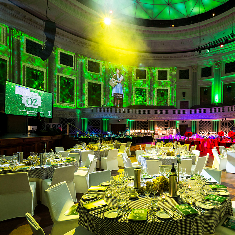 A grand event hall in Brisbane, decorated with green lights and Wizard of Oz projections, showcases round tables set for dinner—all expertly managed by a leading event production Brisbane team.
