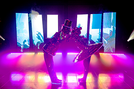Two performers in shiny, reflective costumes and hats are posing face-to-face under colorful stage lights, with bright LED screens displaying abstract visuals behind them, creating an unforgettable encore moment.