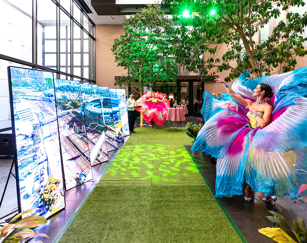 Two dancers in colorful, butterfly-inspired costumes perform beside large digital screens and indoor trees. The setting features a green carpet, plants, and vibrant, festive lighting.