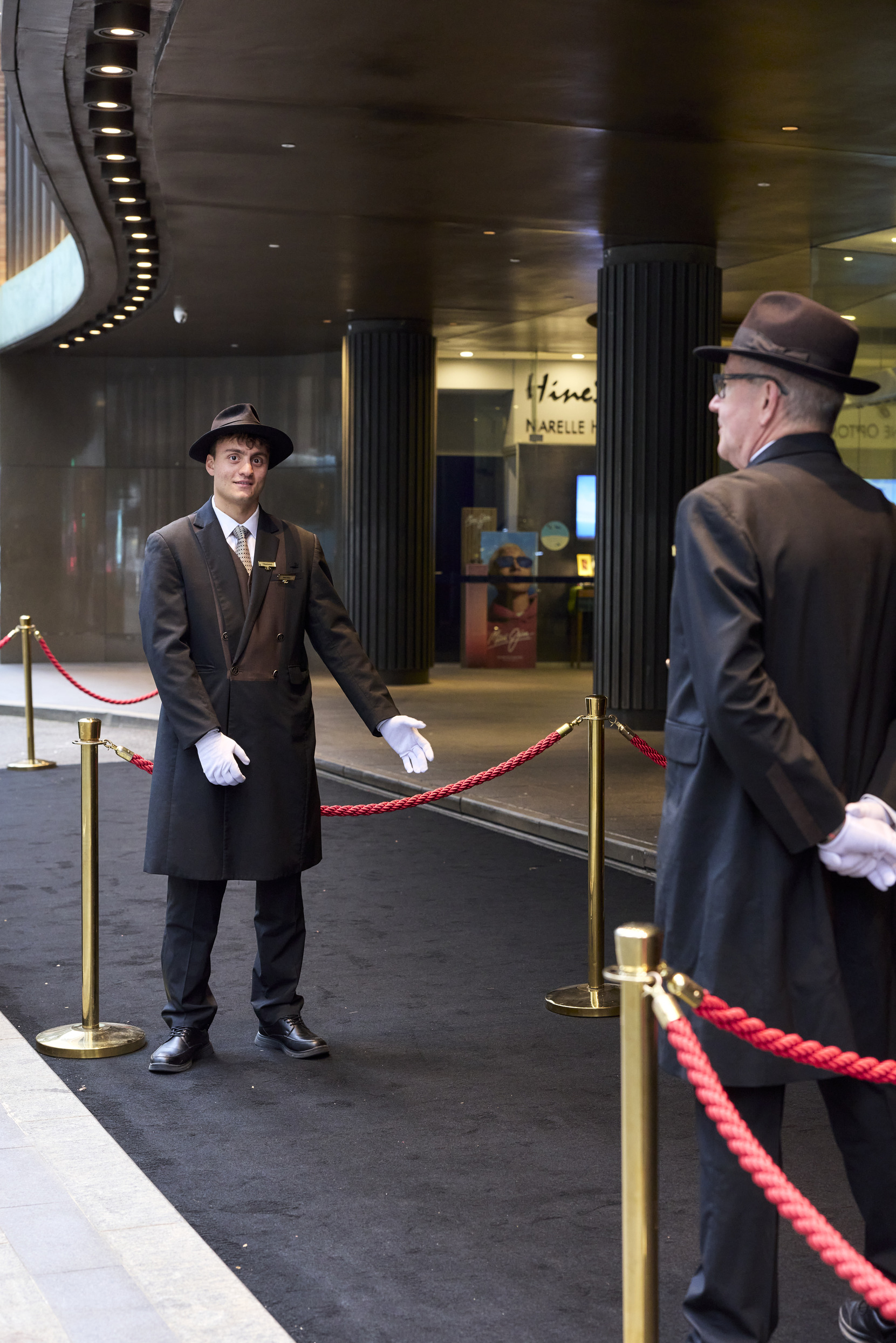 Two doormen in black suits, hats, and white gloves stand on either side of a roped-off entrance with black carpet outside a building, one gesturing to welcome guests.