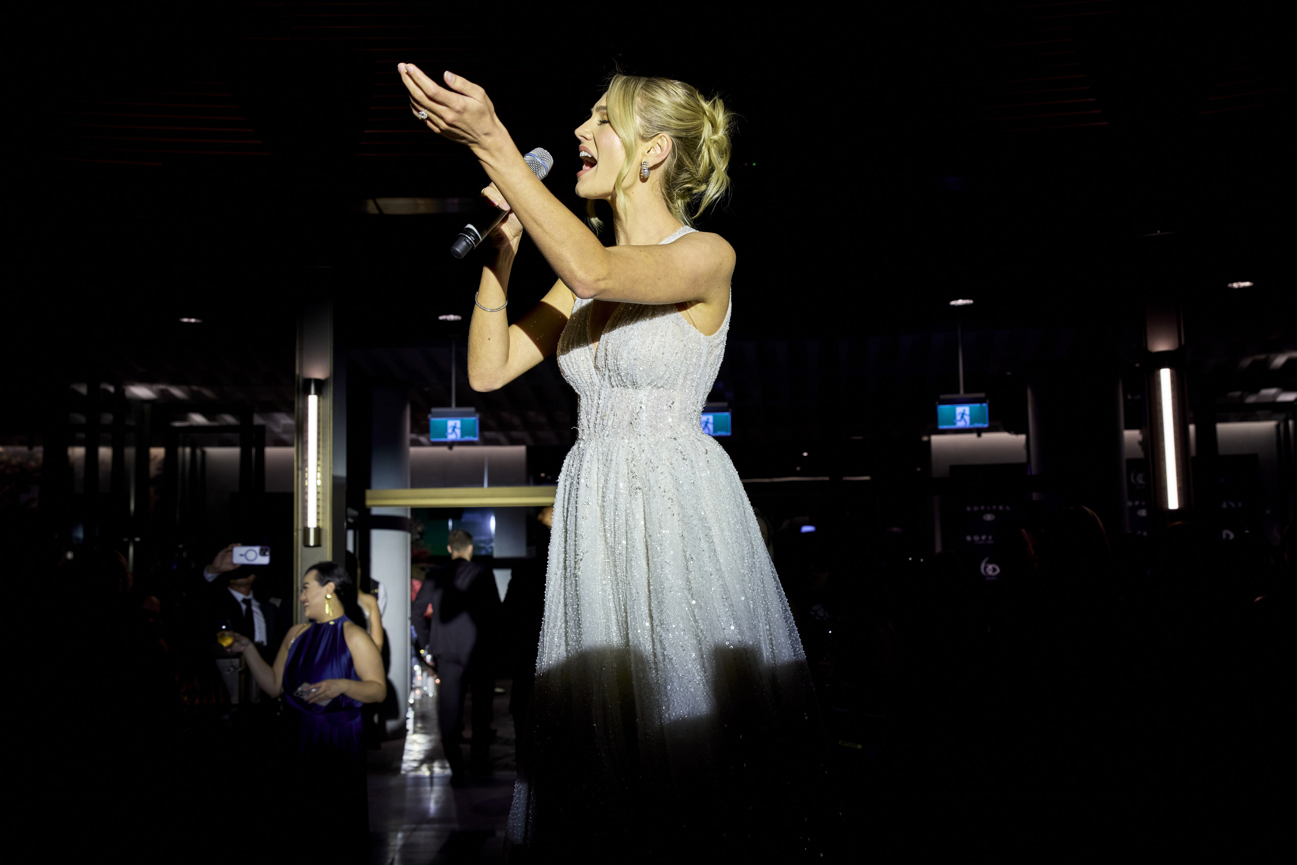 A woman in a shimmering white gown sings passionately into a microphone under a spotlight, surrounded by darkness and an audience in a dimly lit venue.