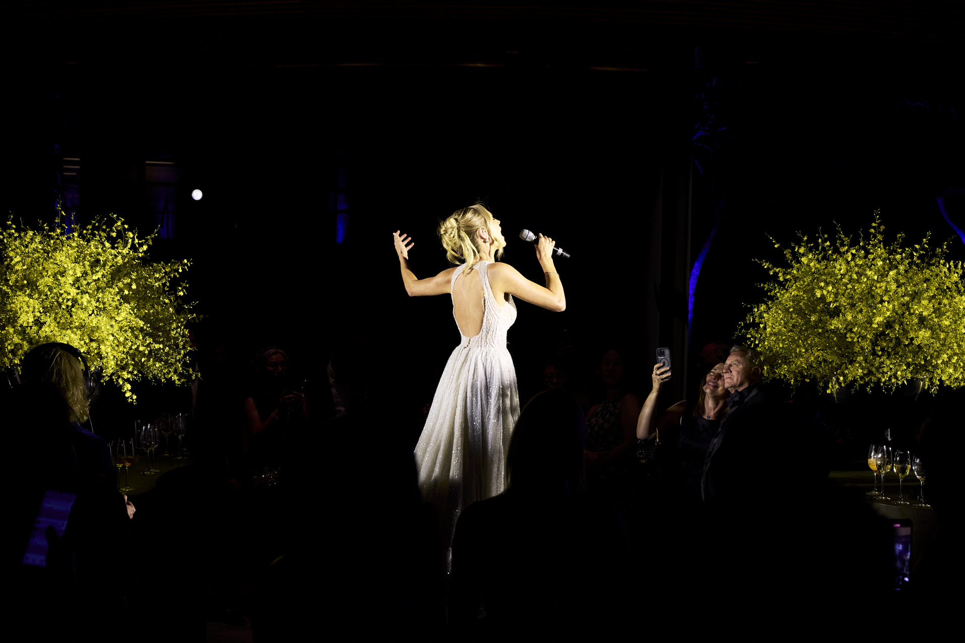 A woman in a sparkling, backless gown sings into a microphone on a dimly lit stage, illuminated by spotlights, with yellow floral arrangements glowing behind her and an audience watching.
