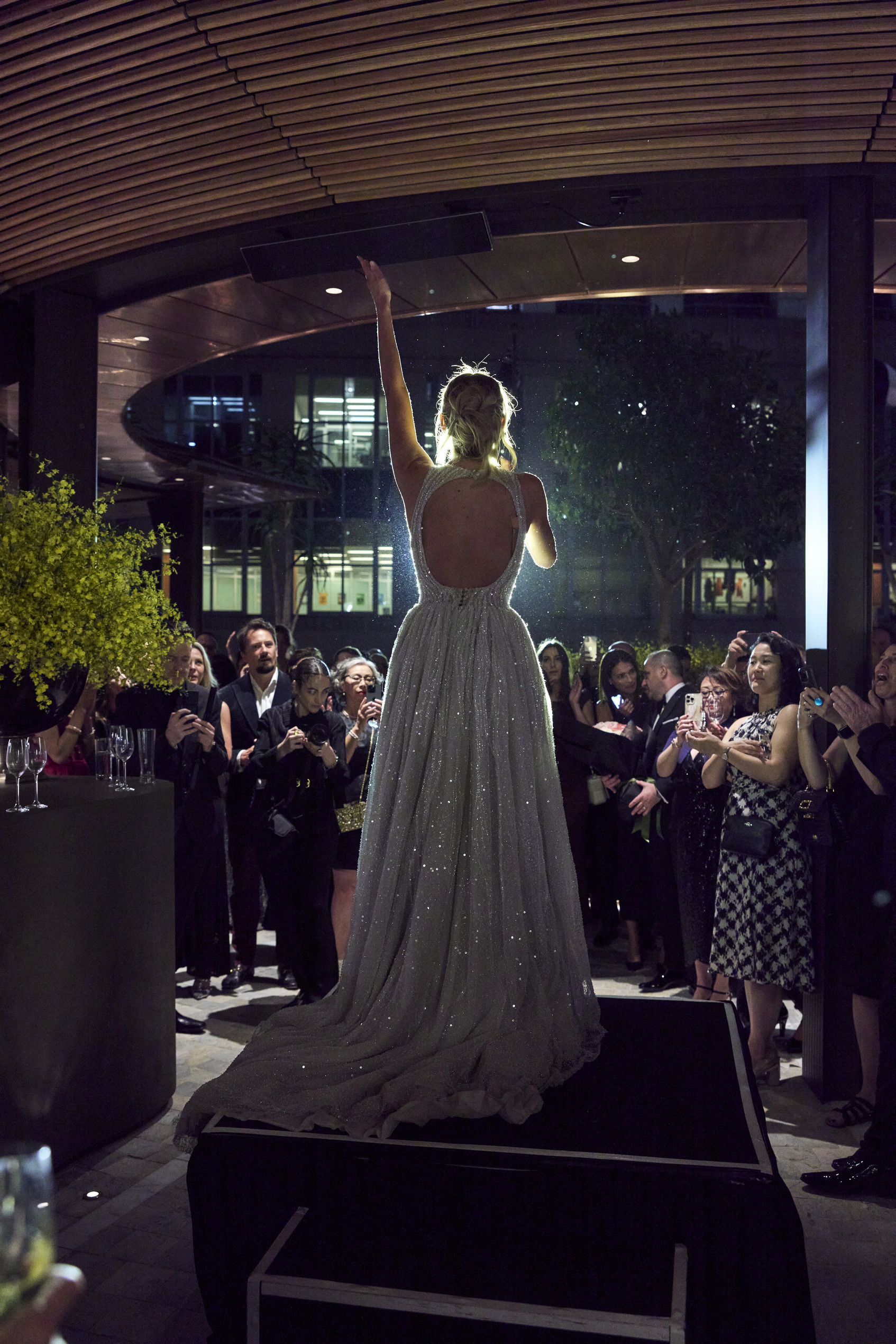 A woman in a sparkling, backless gown stands on a platform, raising one arm as she addresses a crowd of people dressed in formal attire at an evening event.