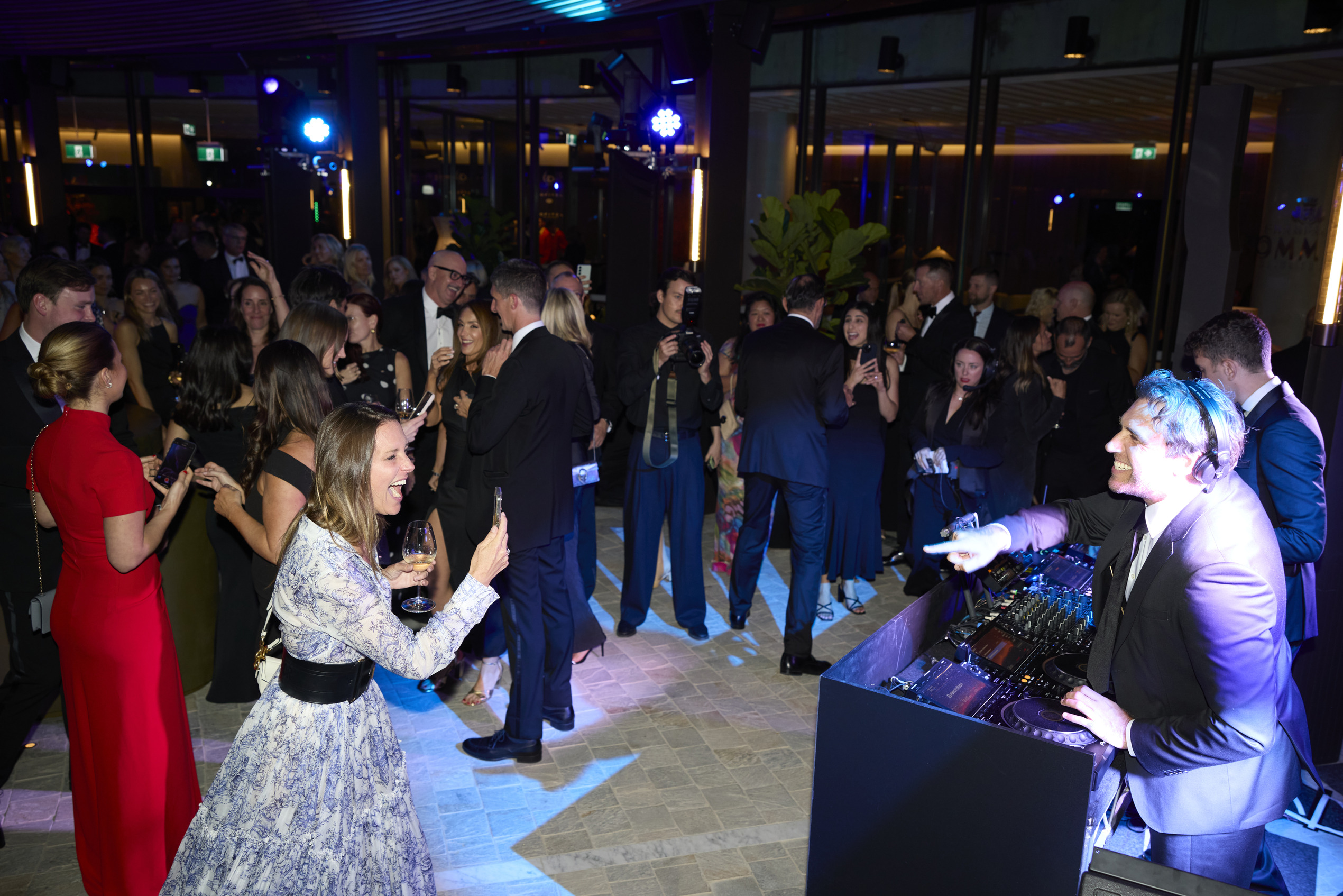 A lively crowd in formal attire enjoys a party indoors, with people dancing and taking photos. A DJ with headphones is performing at a booth under bright lights. The atmosphere is energetic and festive.