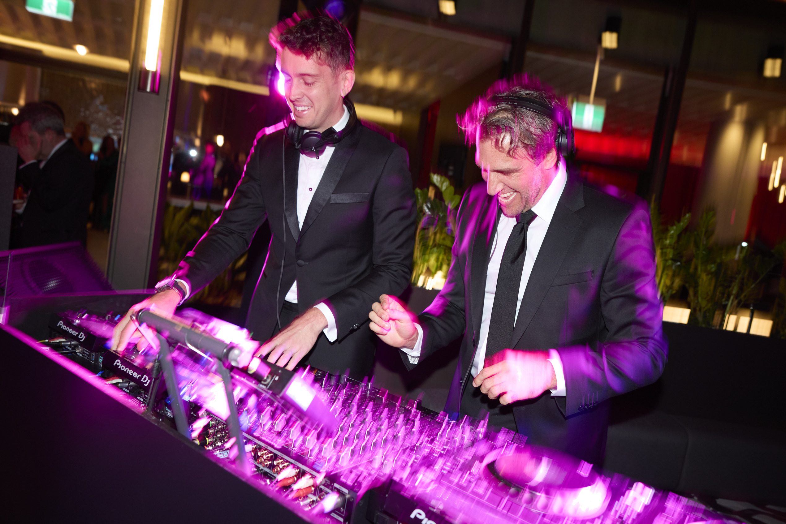 Two men in tuxedos and headphones laugh and DJ together at a brightly lit indoor event, surrounded by DJ equipment and colorful lights.