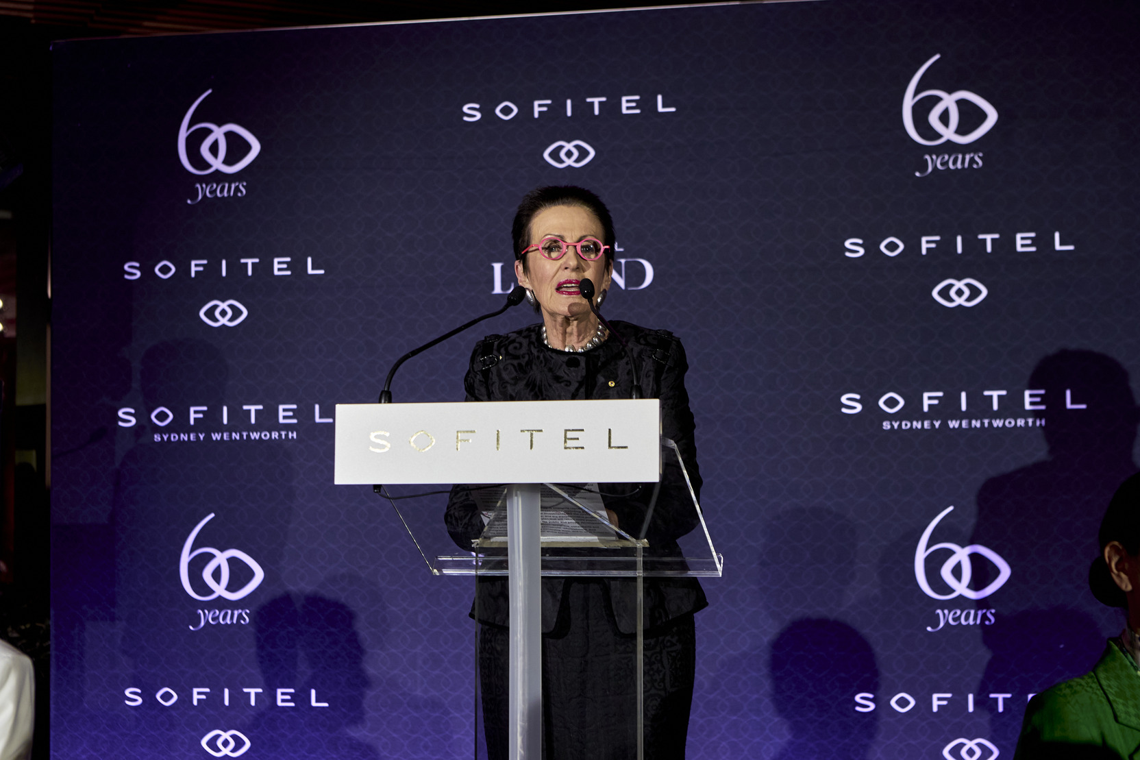 A woman wearing glasses and a black outfit speaks at a podium with a Sofitel sign, against a backdrop displaying Sofitel, 60 years, and Sydney Wentworth.