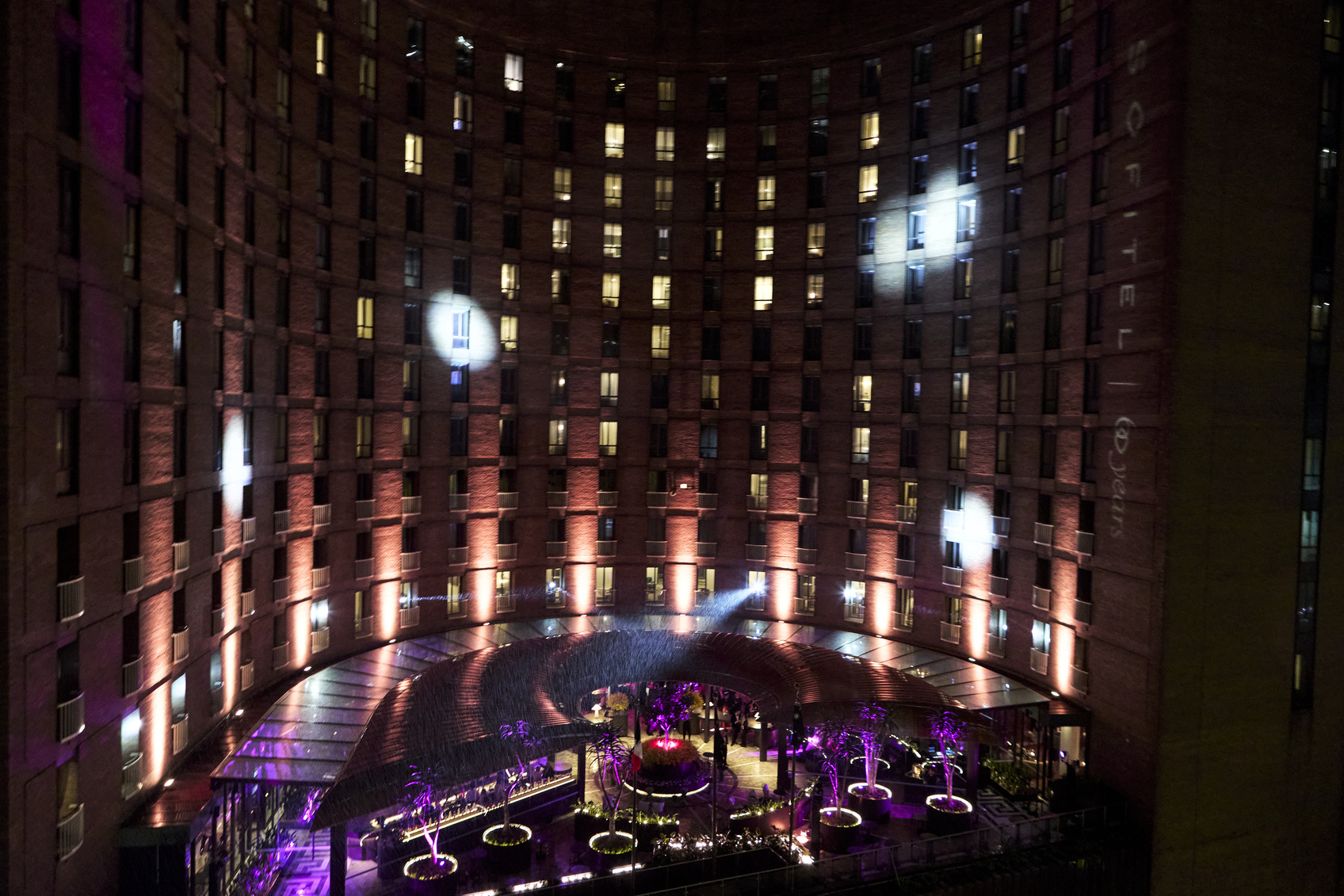 A nighttime view of a circular hotel courtyard lit with purple and white lights, surrounded by tall, curved brick walls with many windows. The central area features illuminated trees and seating.