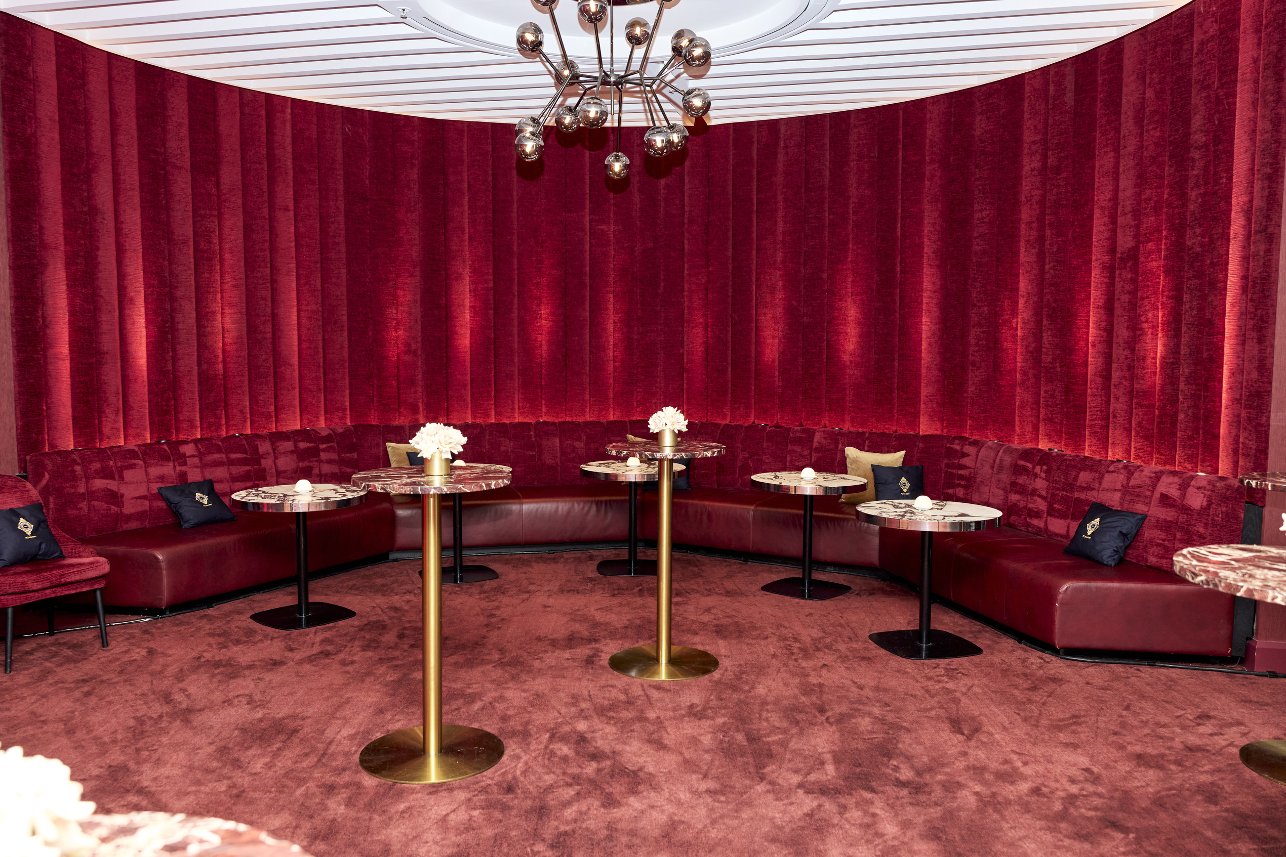 A luxurious lounge with red velvet curved seating, round marble tables with gold bases, small floral centerpieces, and plush cushions, set against red velvet walls and carpet. A modern chandelier hangs from the ceiling.