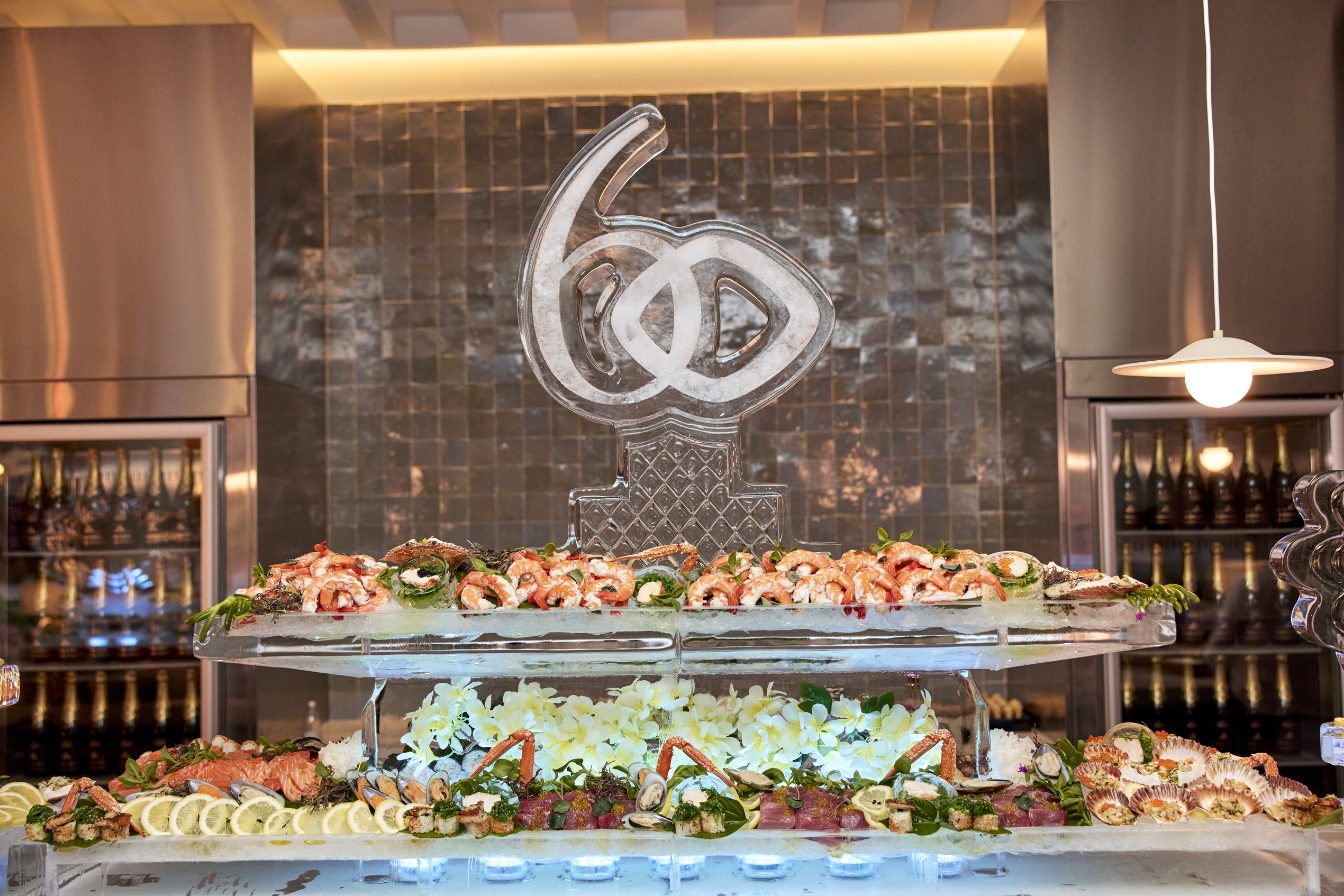 A seafood display with sushi, crab, and shellfish is arranged on ice beneath a decorative ice sculpture shaped like interlocking rings and the number 60, in a modern setting with tile walls and wine bottles.