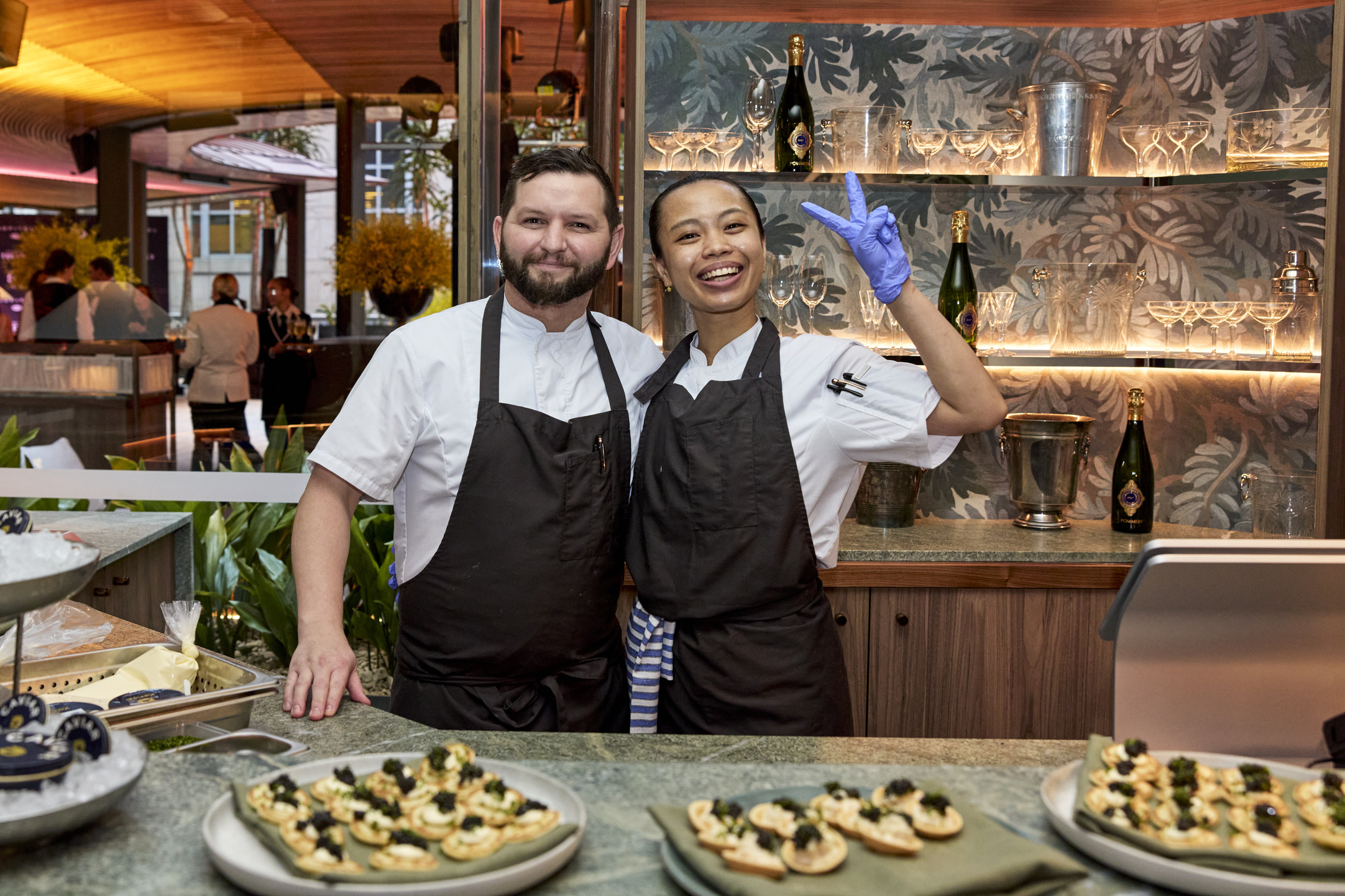 Two smiling chefs in black aprons stand behind a counter with plates of appetizers. One chef has their arm around the other, who is wearing a glove and flashing a peace sign. Bottles and glasses are on the shelves behind them.
