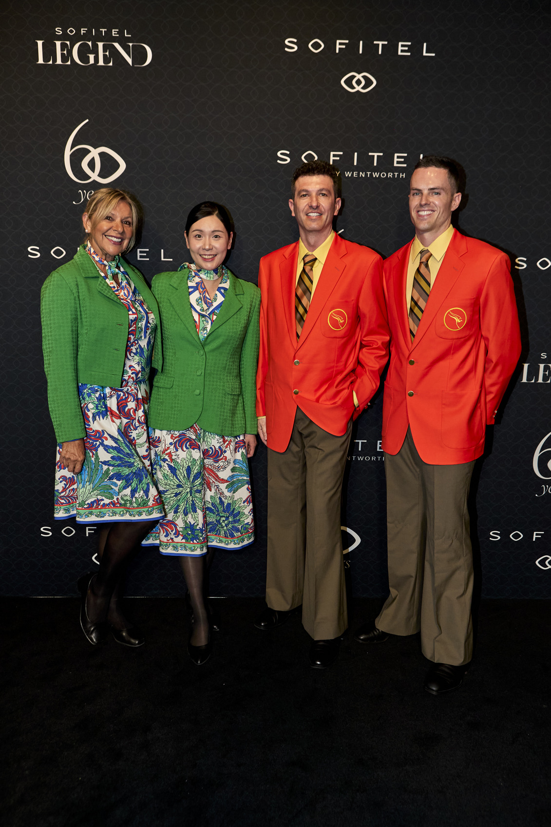 Four people stand on a black carpet in front of a Sofitel Legend backdrop. Two women wear bright patterned dresses with green blazers, and two men wear orange blazers, striped ties, and tan pants.