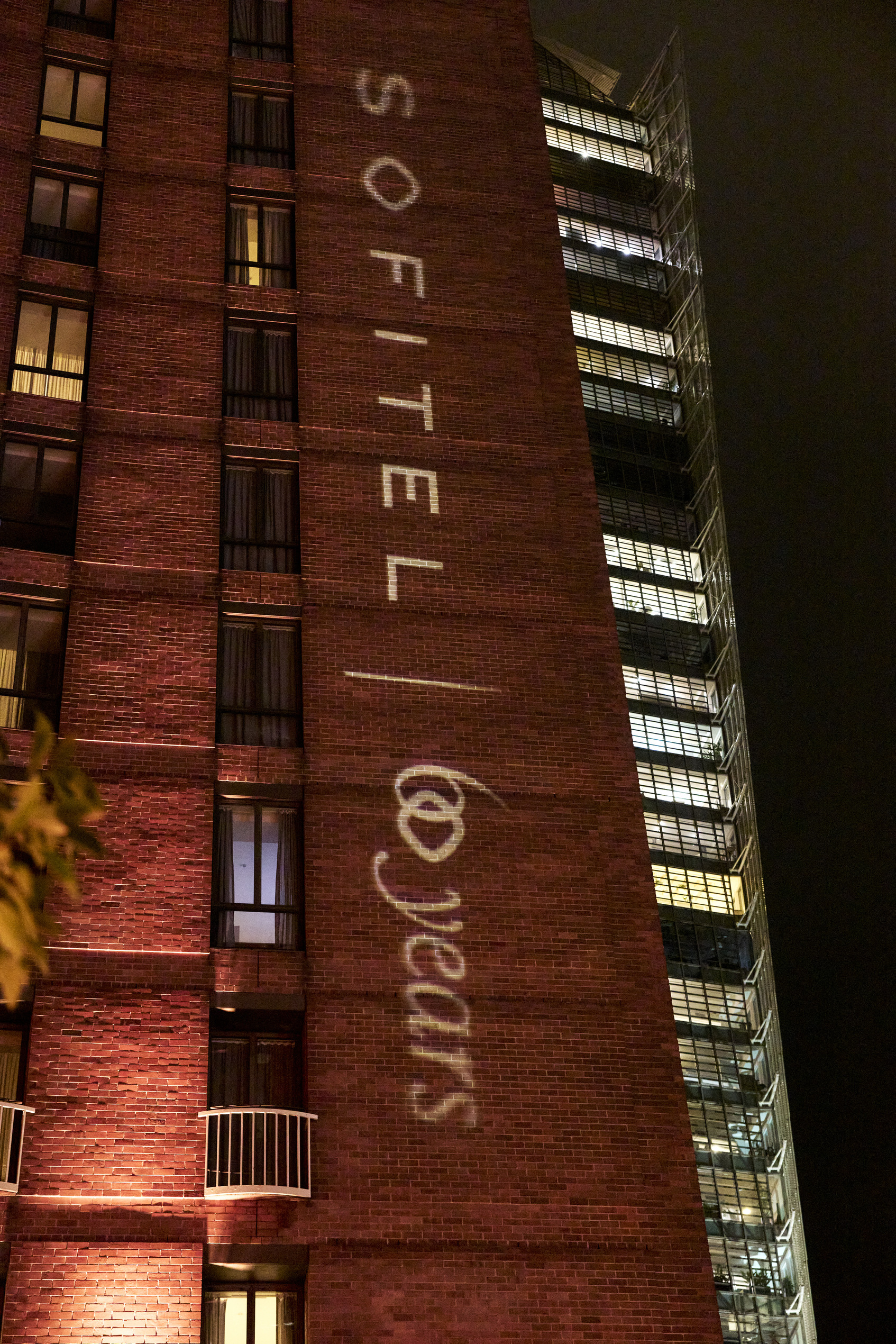 A nighttime view of a red brick hotel building with the words “SOFITEL 60 years” illuminated vertically in white light on its exterior wall. Some windows are lit, and a modern glass building is visible in the background.