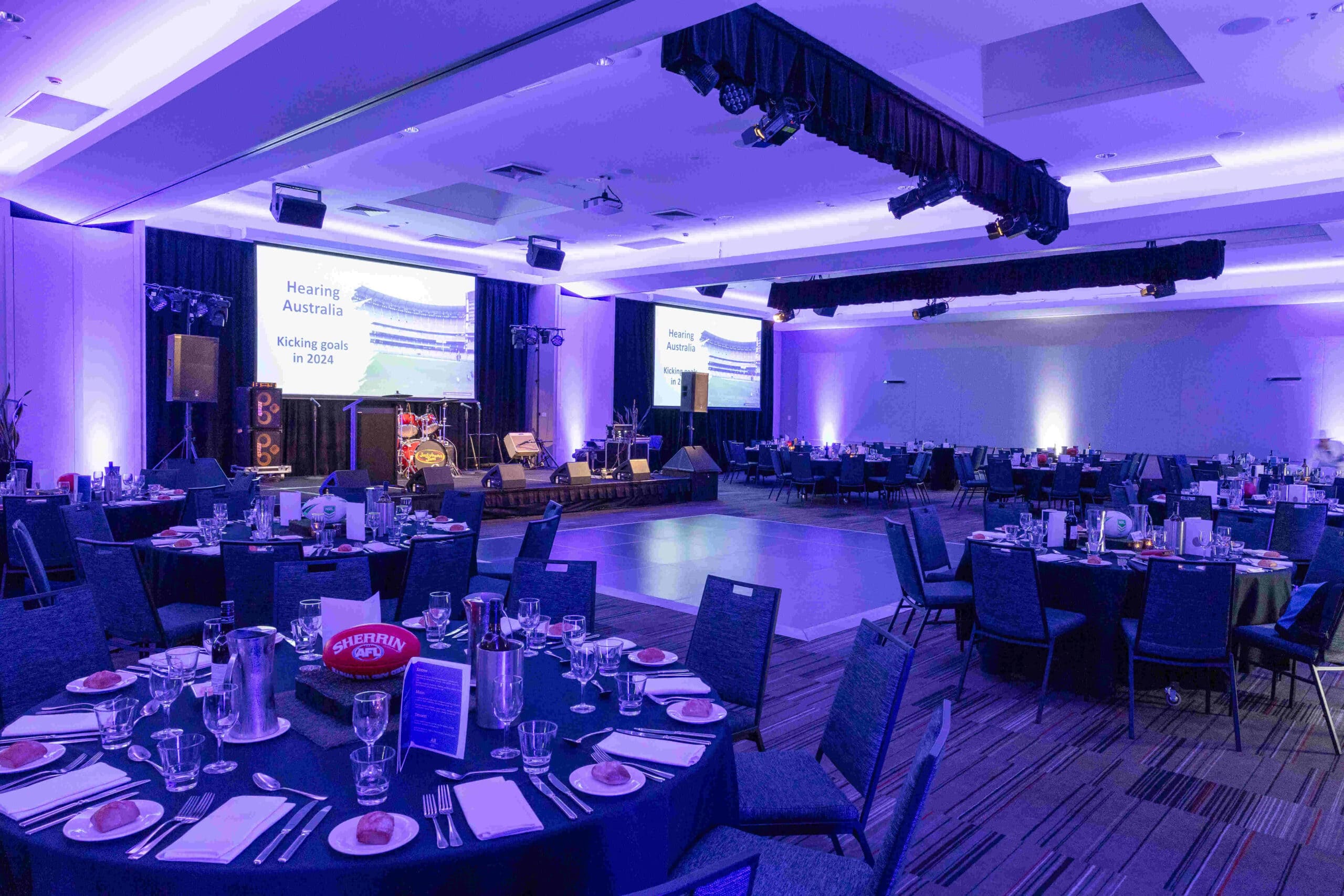 A large banquet hall set up for an event, with round tables arranged around a central dance floor, formal place settings, blue-purple lighting, and projection screens displaying “Hearing Australia Kicking goals in 2023.”.