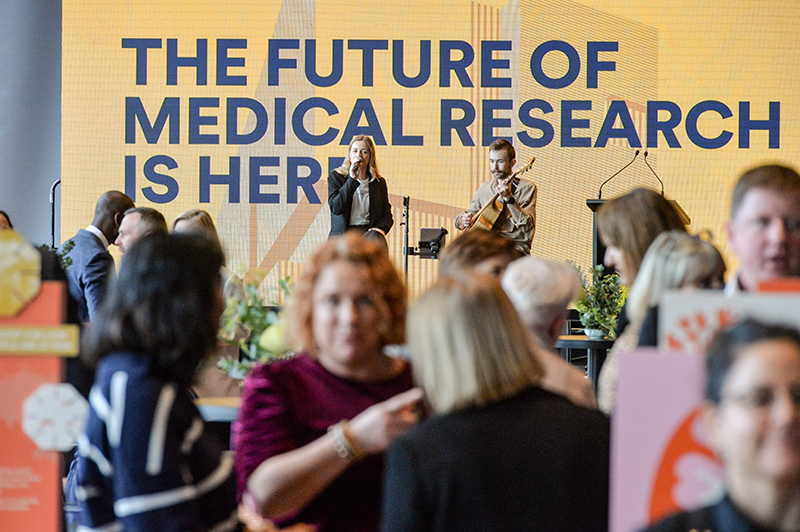 A crowd gathers at an event with a large yellow sign in the background reading The Future of Medical Research Is Here. Two people stand on stage, one speaking at a podium and the other standing nearby.