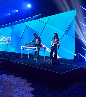 Two musicians perform on a stage with bright blue geometric LED screens powered by event technology at an indoor event. One plays a keyboard or DJ controller, while the other rocks out on an electric guitar.