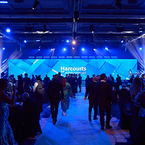 People in formal attire gather in a large, blue-lit event hall with a stage at the front displaying the Harcourts logo. The atmosphere is lively as attendees walk, mingle, and anticipate an encore of exciting moments.
