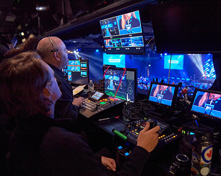Two people operate cameras and monitors in a control booth at a live Encore event, watching various video feeds of a stage with blue lighting and a speaker addressing the audience.