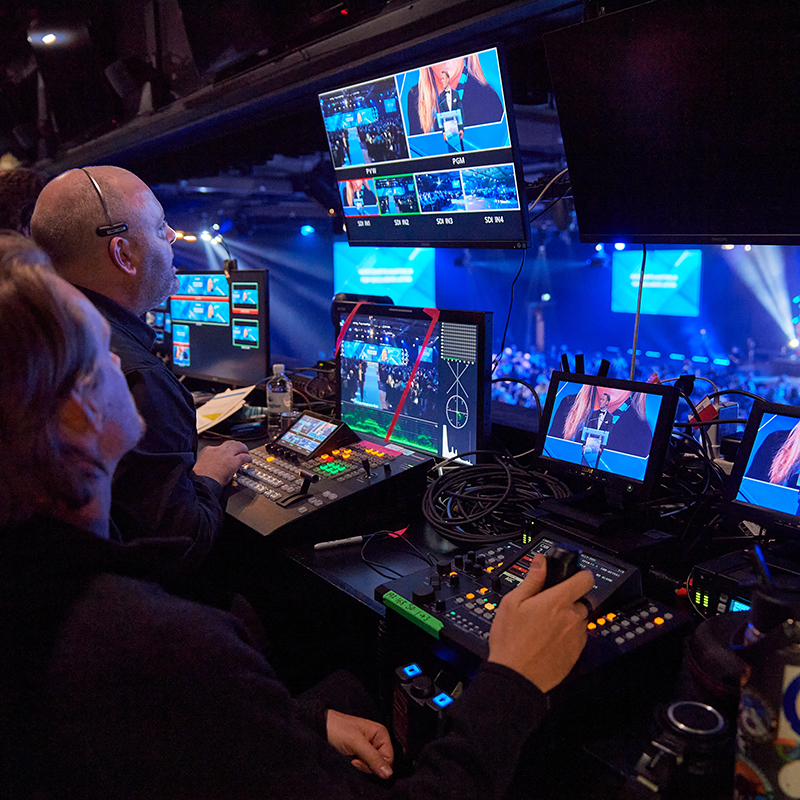 Two people operate video and audio equipment in a control booth, monitoring multiple screens showing a stage event with a large audience in the background, under blue and purple lighting.