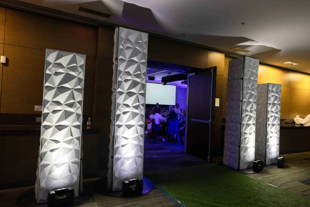 Four tall, geometric-patterned white columns are illuminated at the entrance of a dimly lit event space, where people are visible inside seated at tables under blue lighting.